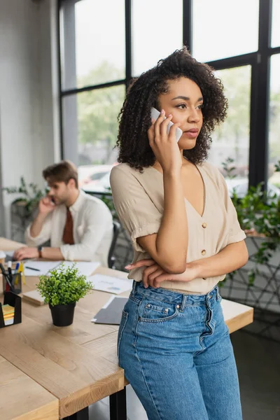 Ernsthafte junge afrikanisch-amerikanische Frau telefoniert in der Nähe eines verschwommenen Kollegen, der im modernen Büro auf dem Smartphone spricht — Stockfoto