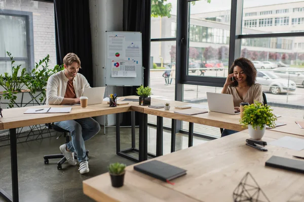 Lächelnde junge afrikanisch-amerikanische Frau telefoniert in der Nähe ihres Kollegen, der im modernen Büro am Laptop arbeitet — Stockfoto