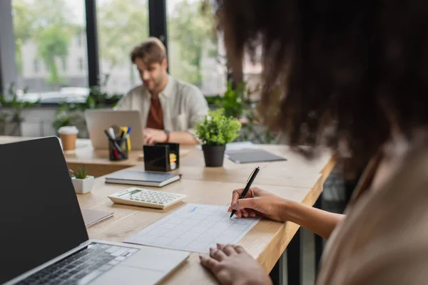 Partial view of young african american woman writing with pen near colleague sitting with laptop in modern office — Stock Photo