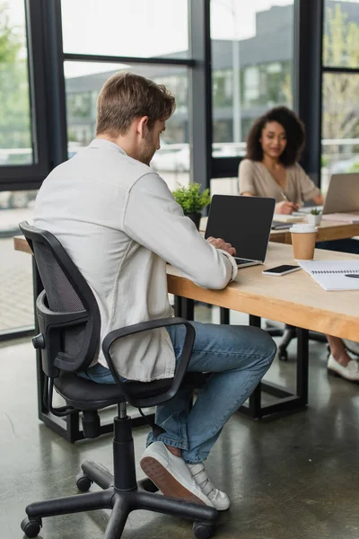 Interracial colleagues sitting at desks and working with laptops in modern open space — Stock Photo