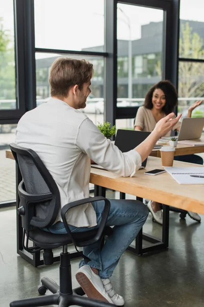 Interracial colleagues sitting at desks near laptops and gesturing with hands in modern open space — Stock Photo