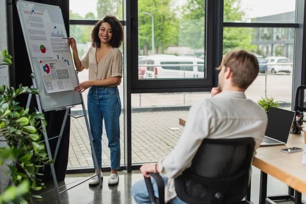 Sonriente joven afroamericana mujer de pie cerca de rotafolio y hacer la presentación a su colega en la oficina - foto de stock