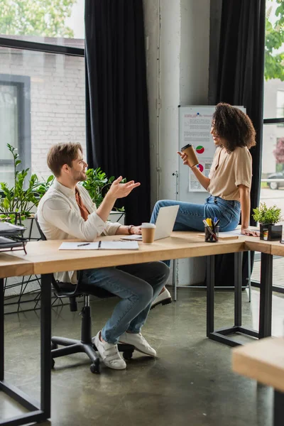 Giovane donna afroamericana sorridente con tazza di carta seduta sulla scrivania vicino al collega con computer portatile in ufficio moderno — Foto stock