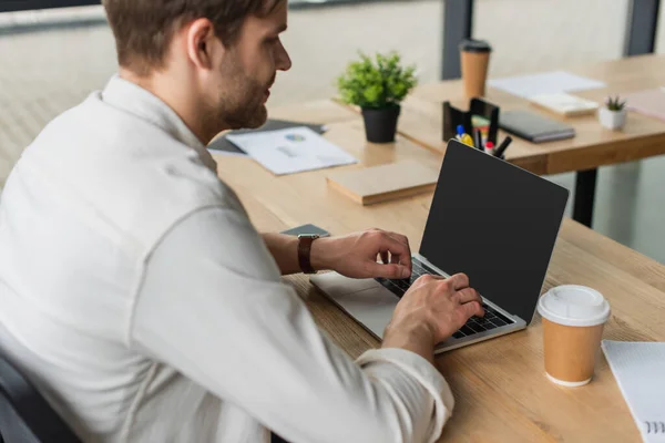 Молодой человек сидит за столом и печатает на ноутбуке с чистым экраном в современном офисе — стоковое фото