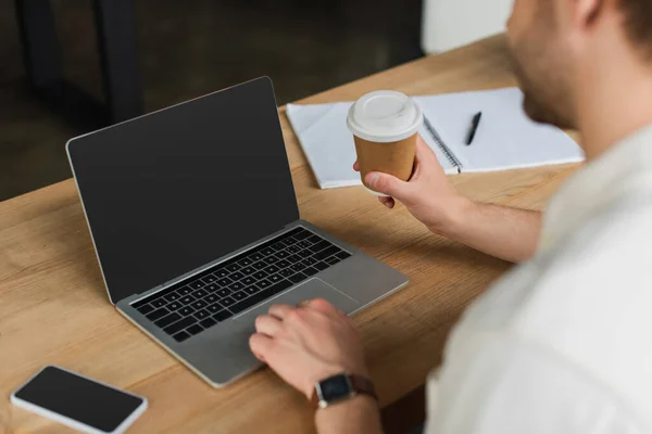 Vista parcial del joven sentado con la taza de papel en la mano y escribir en el ordenador portátil con pantalla en blanco en la oficina moderna - foto de stock