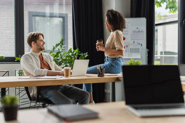 Junge afrikanisch-amerikanische Frau mit Pappbecher sitzt auf Schreibtisch neben lächelndem Kollegen mit Laptop im modernen Büro — Stockfoto