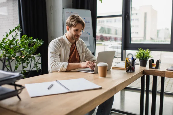 Sorrindo jovem sentado na mesa com copo de papel e digitando no laptop no escritório moderno — Fotografia de Stock