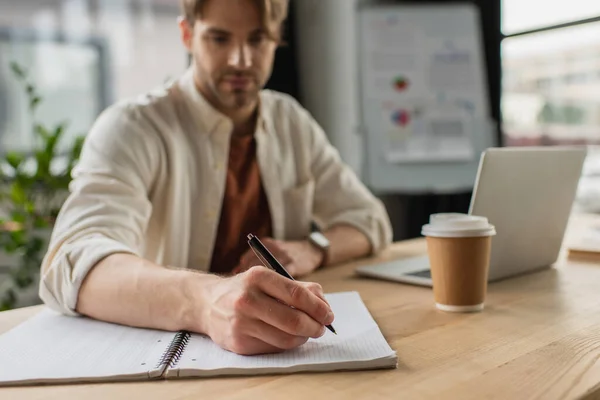 Giovane seduto alla scrivania vicino a tazza di carta e laptop e scrivere con penna nel taccuino in ufficio moderno — Foto stock