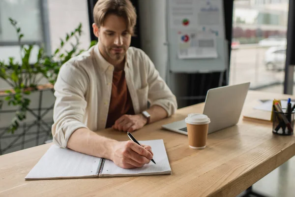 Ernster junger Mann sitzt am Schreibtisch neben Pappbecher und Laptop und schreibt mit Stift in Notizbuch in modernem Büro — Stockfoto