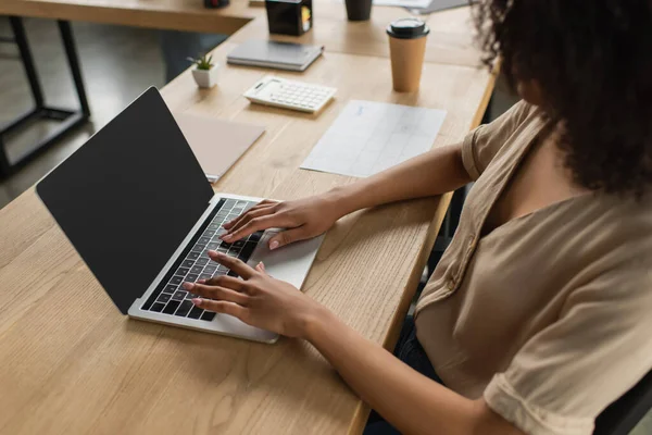 Visão parcial da jovem afro-americana sentada à mesa com xícara de papel e digitando no laptop no escritório moderno — Fotografia de Stock