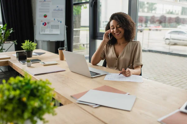 Sorridente giovane donna afroamericana che parla sul cellulare e tiene la matita vicino al computer portatile in ufficio moderno — Foto stock