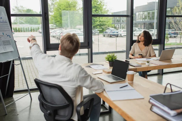 Junge afrikanisch-amerikanische Frau sitzt neben verschwommenem Kollegen und zeigt mit dem Finger auf Flipchart in modernem Büro — Stockfoto