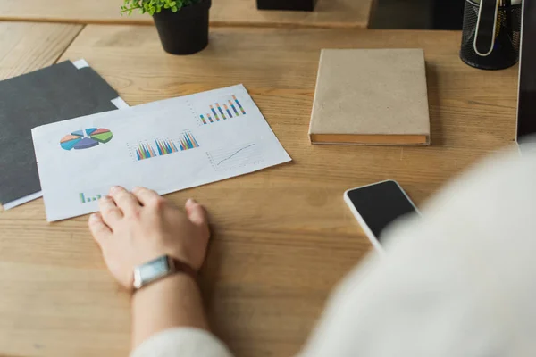 Vista parcial de la mano masculina en papel de toque reloj de pulsera con gráficos y gráficos cerca del teléfono celular en escritorio de madera - foto de stock