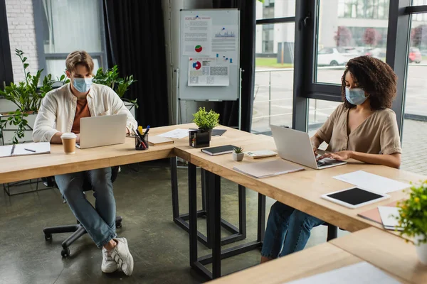 Giovani colleghi interrazziali in maschere mediche che lavorano con i computer portatili in un moderno spazio aperto — Stock Photo