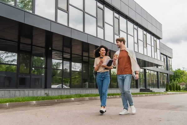 Smiling young interracial couple with paper coffee cups and cellphones in hands walking on street near modern building — Stock Photo