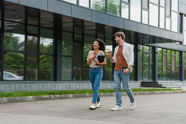 Positive young interracial couple with paper coffee cups and cellphones in hands walking on street near modern building — Stock Photo