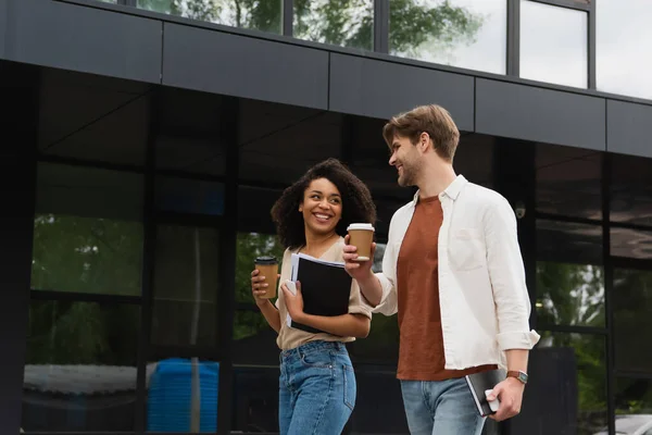 Sorrindo jovem casal interracial com copos de papel e celulares em mãos olhando uns para os outros e andando perto do edifício — Fotografia de Stock