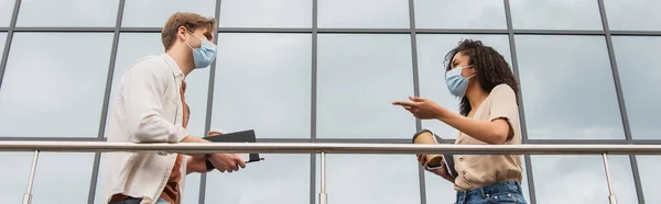 Низкий угол обзора молодых межрасовых пар в медицинских масках разговаривающих возле стеклянного здания на улице, баннер — стоковое фото