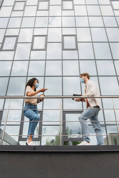 Low angle view of young interracial couple in medical masks and casual clothes talking near glass building on street — Stock Photo