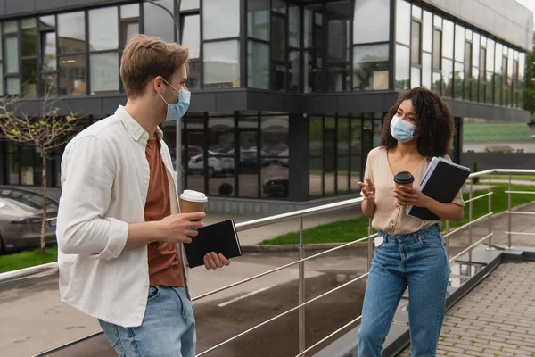 Giovane coppia interrazziale in maschere mediche e vestiti casual parlando sulla strada vicino edificio di vetro — Foto stock