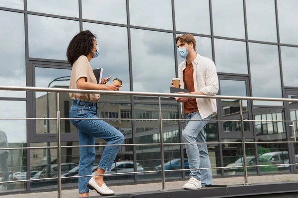 Young interracial couple in medical masks and casual clothes talking with paper cups in hands near glass building on street — Stock Photo