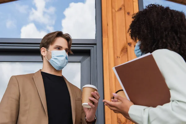 Giovani colleghi interrazziale in maschere mediche parlando con bicchieri di carta in mano vicino edificio sulla strada — Stock Photo