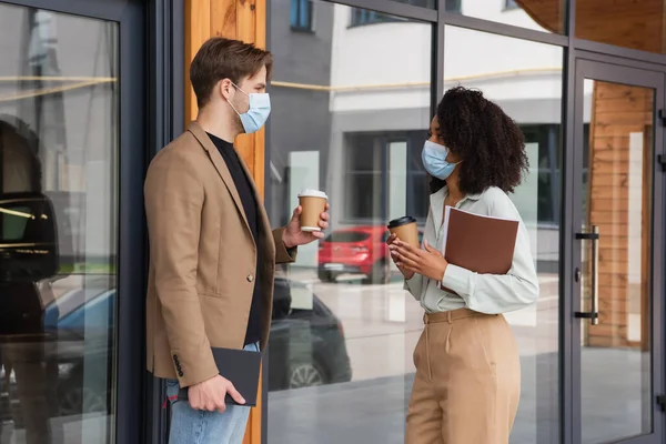 Jóvenes colegas interracial en máscaras médicas hablando con vasos de papel en las manos en la calle cerca del edificio - foto de stock