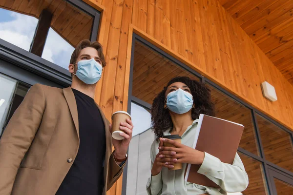 Low angle view of young interracial colleagues in medical masks standing with paper cups in hands on street near building — Stock Photo