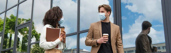 Молоді міжрасові колеги в медичних масках, що ходять з паперовими стаканчиками та документами біля скляної будівлі, банер — стокове фото
