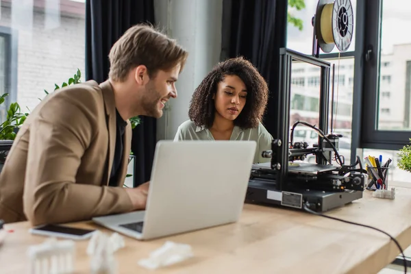 Lächelnder junger Mann sitzt mit afrikanisch-amerikanischem Kollegen in der Nähe von 3D-Drucker und Laptop im modernen Büro — Stockfoto