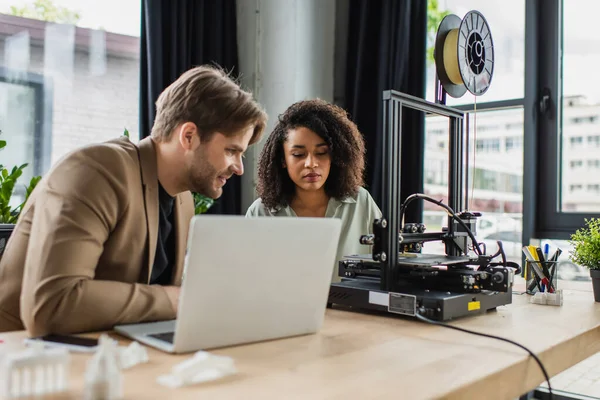 Colegas interraciales sentados cerca de la impresora 3D, portátil y figuras de plástico en la oficina moderna - foto de stock