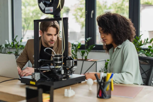 Serious interracial colleagues sitting near 3D printer, laptop and plastic figures in modern office — Stock Photo