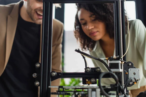 Jóvenes colegas interracial mirando a la impresora 3D produciendo figura de plástico en la oficina moderna - foto de stock