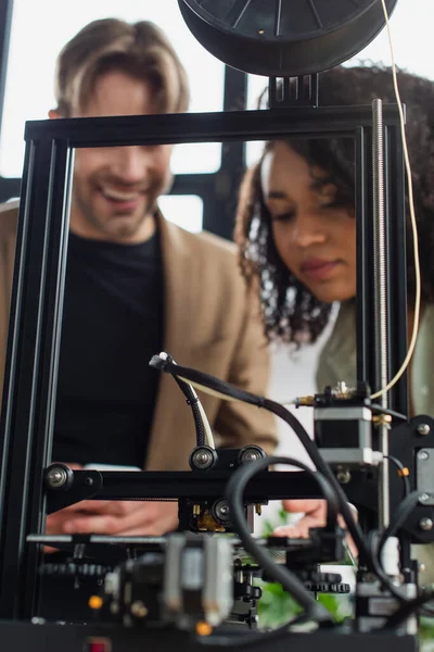 Impresora 3D produciendo figura de plástico cerca borrosa jóvenes colegas interracial en el fondo - foto de stock