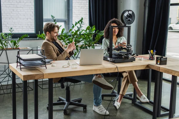 Lächelnde Kollegen, die in der Nähe von 3D-Drucker und Laptop im modernen Büro auf Handys Nachrichten senden — Stockfoto