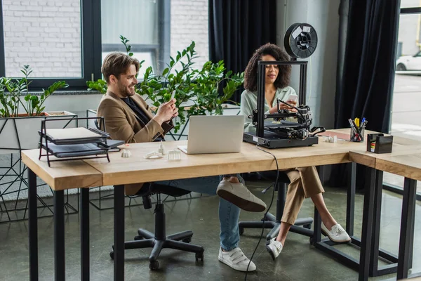 Smiling interracial colleagues messaging on cellphones near 3D printer and laptop in modern office — Stock Photo