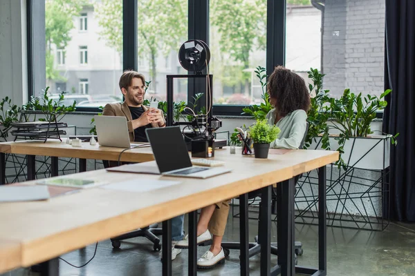 Lächelnde Kollegen mit Handys in der Hand in der Nähe von 3D-Druckern und Laptops im modernen Büro — Stockfoto