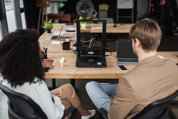 Visão traseira de jovens designers inter-raciais sentados perto de impressora 3D no espaço aberto moderno — Fotografia de Stock