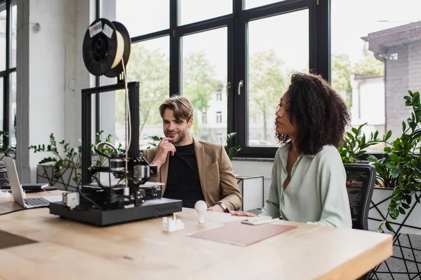 Young interracial designers sitting near 3D printer, plastic figures and laptop in modern open space — Stock Photo