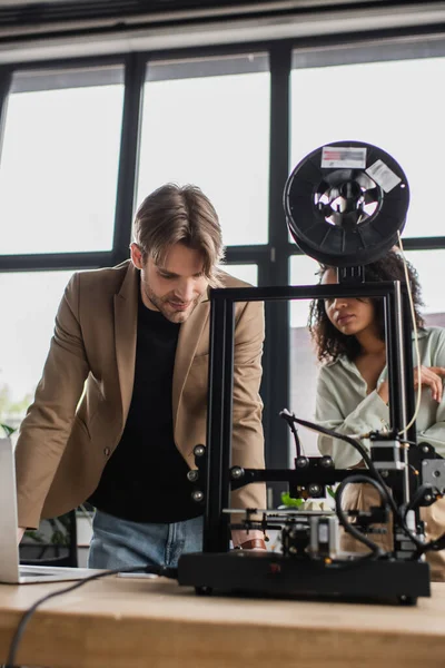 Jóvenes diseñadores interraciales enfocados trabajando con la impresora 3D en la oficina moderna - foto de stock