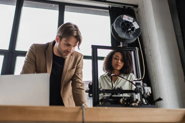 Concentrated young interracial designers working with 3D printer near laptop in modern office — Stock Photo