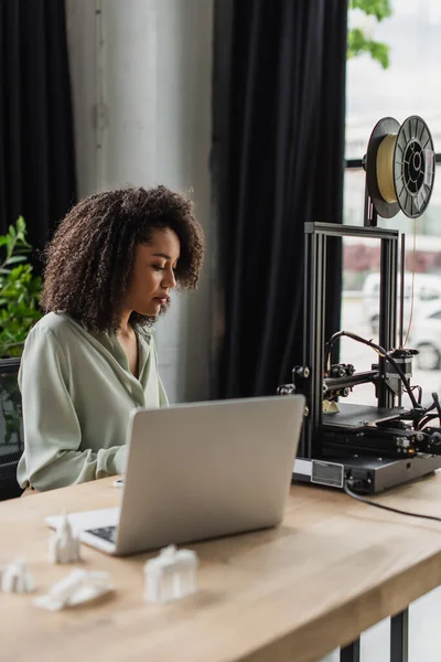 Young african american designer sitting near 3D printer and laptop in modern open space — Stock Photo