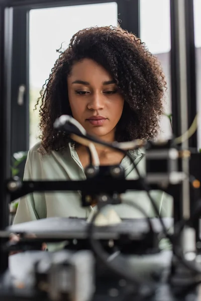 Joven diseñador afroamericano sentado cerca de la impresora 3D en el espacio abierto moderno - foto de stock