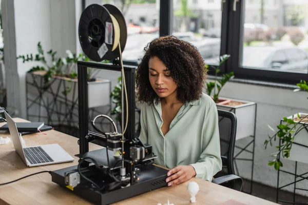 Jovem designer sério trabalhando com impressora 3D perto do laptop e modelos de plástico produzidos na mesa no escritório moderno — Fotografia de Stock