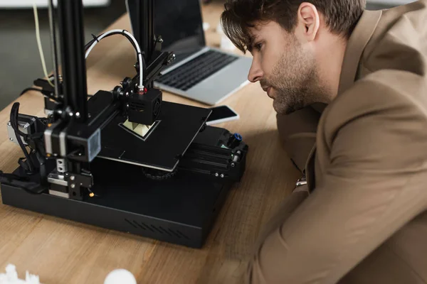 Konzentrierter junger Mann blickt in modernem Büro auf 3D-Drucker mit Plastikfigur — Stockfoto