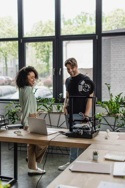 Улыбающиеся межрасовые коллеги, стоящие у стола с ноутбуком и 3D принтером в современном офисе — стоковое фото