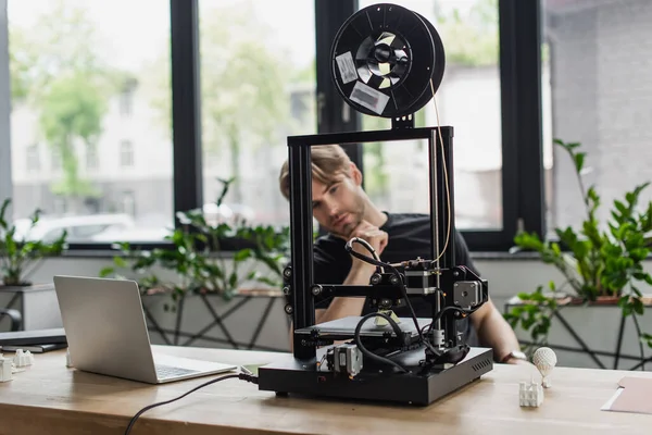 Young designer looking at 3D printer on table with laptop in office — Stock Photo