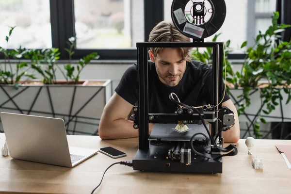 Enfocado joven diseñador sentado cerca de la computadora portátil y mirando a la impresora 3D en la oficina moderna - foto de stock