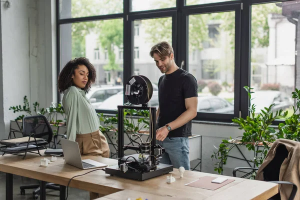 Межрасовые коллеги стоят рядом с 3D принтером, ноутбуком и пластиковыми фигурами в современном офисе — стоковое фото
