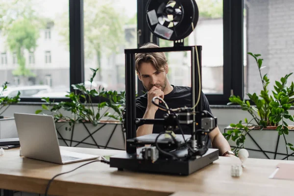 Focused young designer sitting near laptop and plastic figures and looking at 3D printer in modern office — Stock Photo